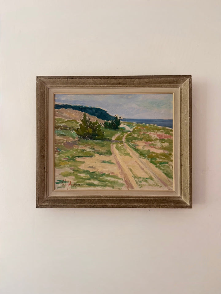 A framed landscape oil painting hanging on a plain off-white wall. The painting depicts a dirt path winding through grassy dunes with small green shrubs towards a distant blue sea under a cloudy sky.