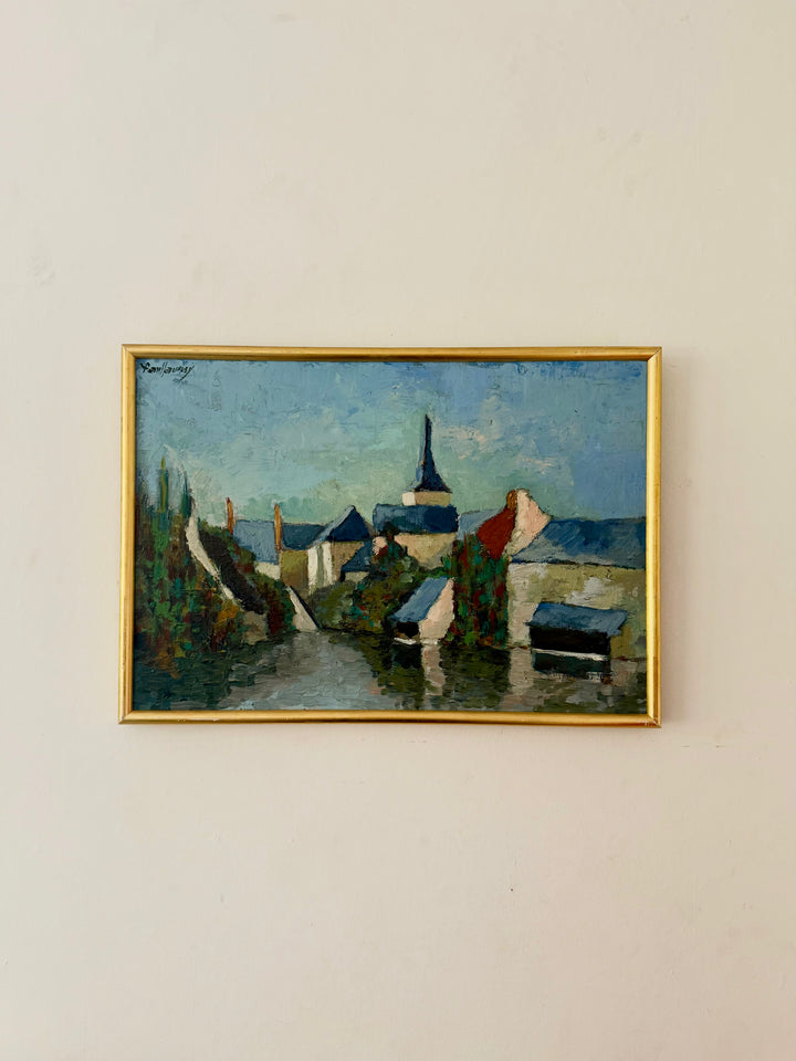 A framed landscape painting of a village with a church spire, hanging on a plain off-white wall. The artwork features blue sky, green trees, and dark buildings, enclosed in a thin gold frame.