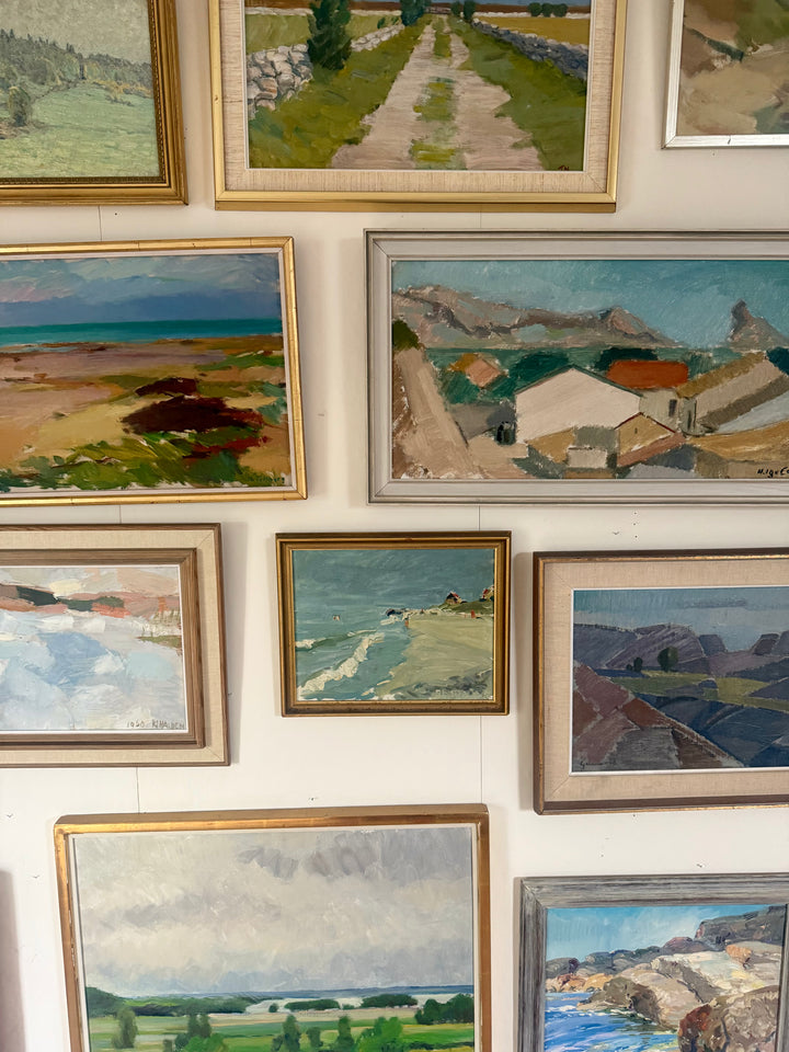A gallery wall display of multiple framed landscape paintings in various gold, silver, and bronze frames, showcasing different coastal and valley scenes.