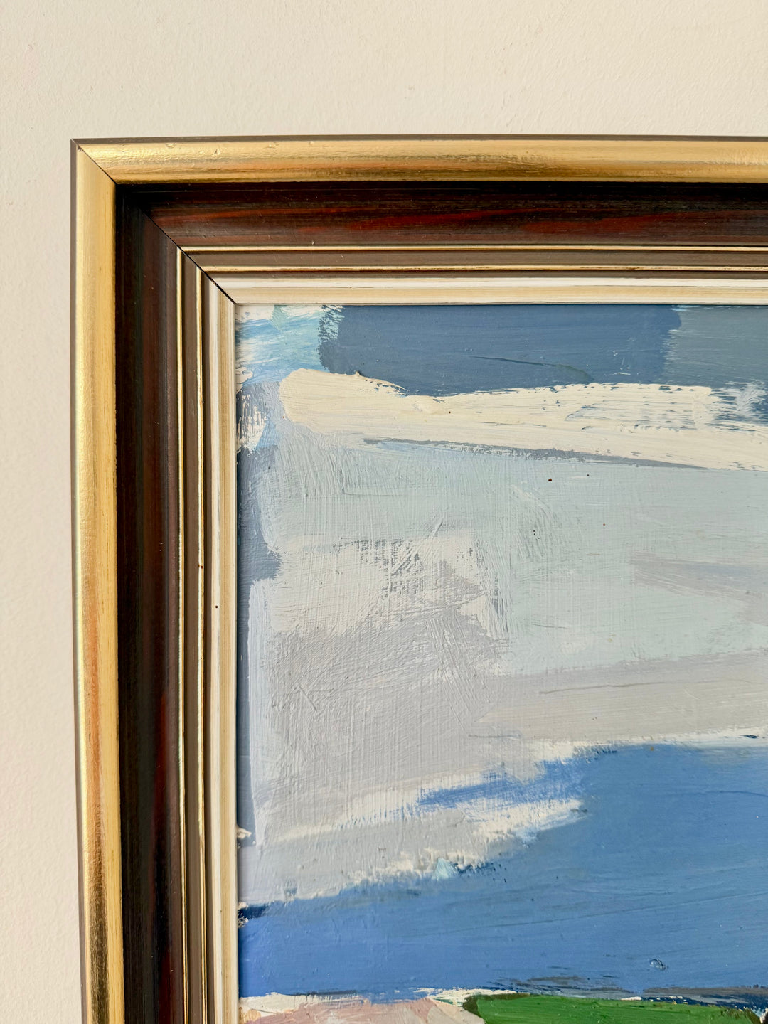 Close-up of a framed painting showing a textured abstract landscape with blue, white, and gray brushstrokes, set in a gold-toned frame against a light beige wall.