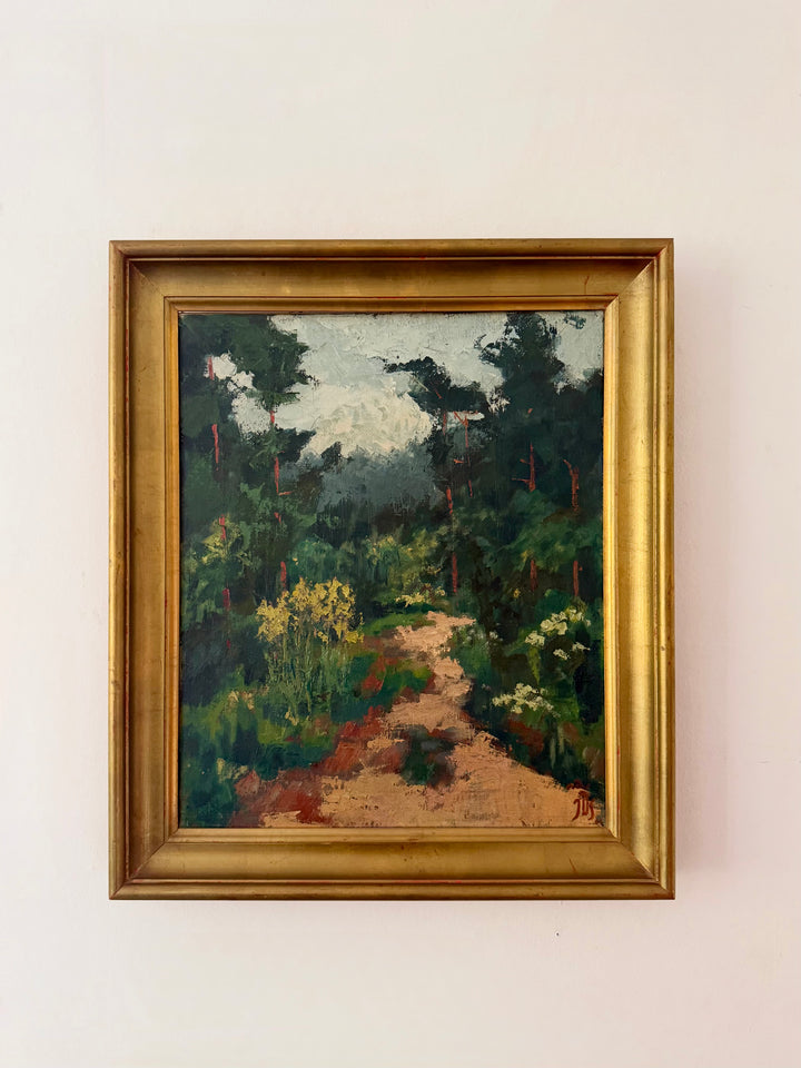A framed landscape oil painting depicting a path through a dense green forest, housed in a wide gold frame and mounted on a plain off-white wall.