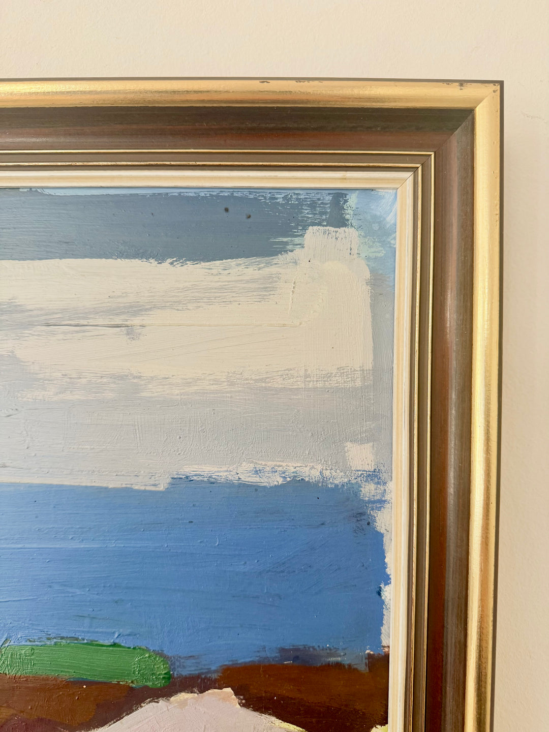A close-up of a framed abstract landscape painting showing textured brushstrokes of blue, white, green, and brown, set in a gold-toned frame against a light beige wall.