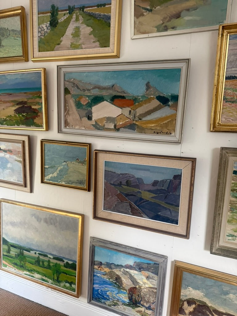 A gallery wall displaying multiple framed landscape paintings in various gold, silver, and brown frames against a white wall.