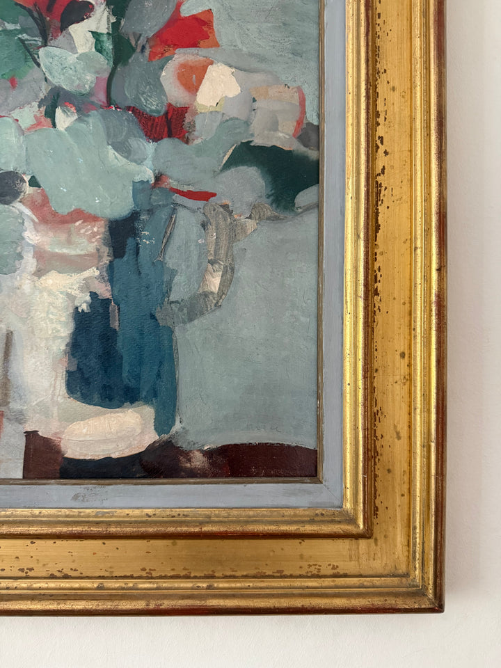Close-up of a framed abstract painting showing textured brushstrokes in blue, gray, white, red, and green, with a distressed gold frame mounted on a light wall.