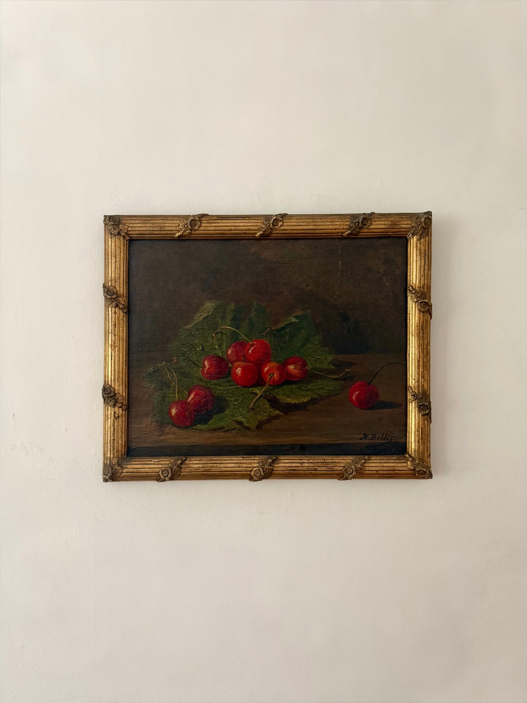 A framed still life painting of red cherries on green leaves, set in a rustic gold-toned wooden frame and mounted on a plain off-white wall.