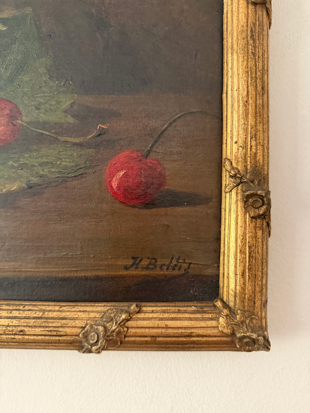 Close-up of a framed still life painting showing red cherries on a dark brown surface, with a distressed gold frame and the artist signature 'A. Belli' visible.