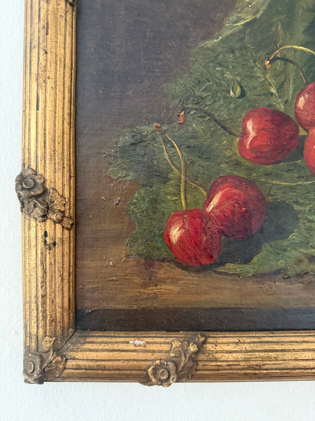 Close-up of a framed still life painting showing red cherries with green leaves on a dark brown background, with an ornate gold-toned frame visible at the left and bottom edges.