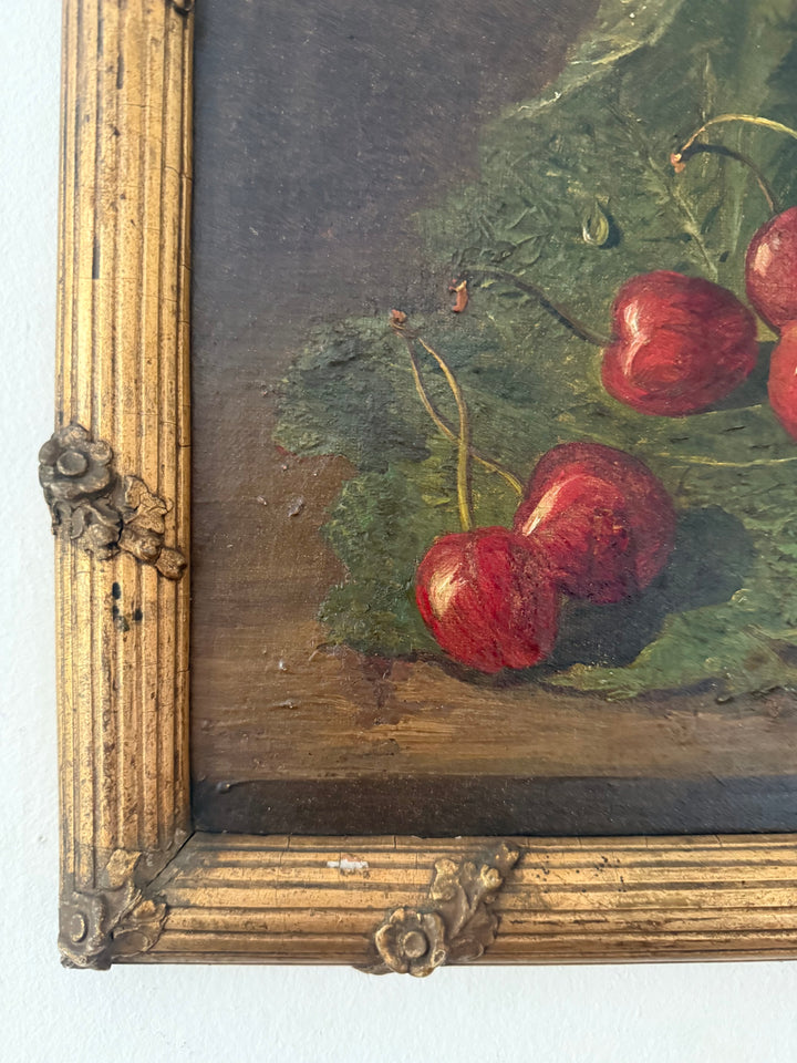 Close-up of a framed still life painting showing red cherries with green leaves on a dark brown background, with an ornate gold-toned frame visible at the left and bottom edges.