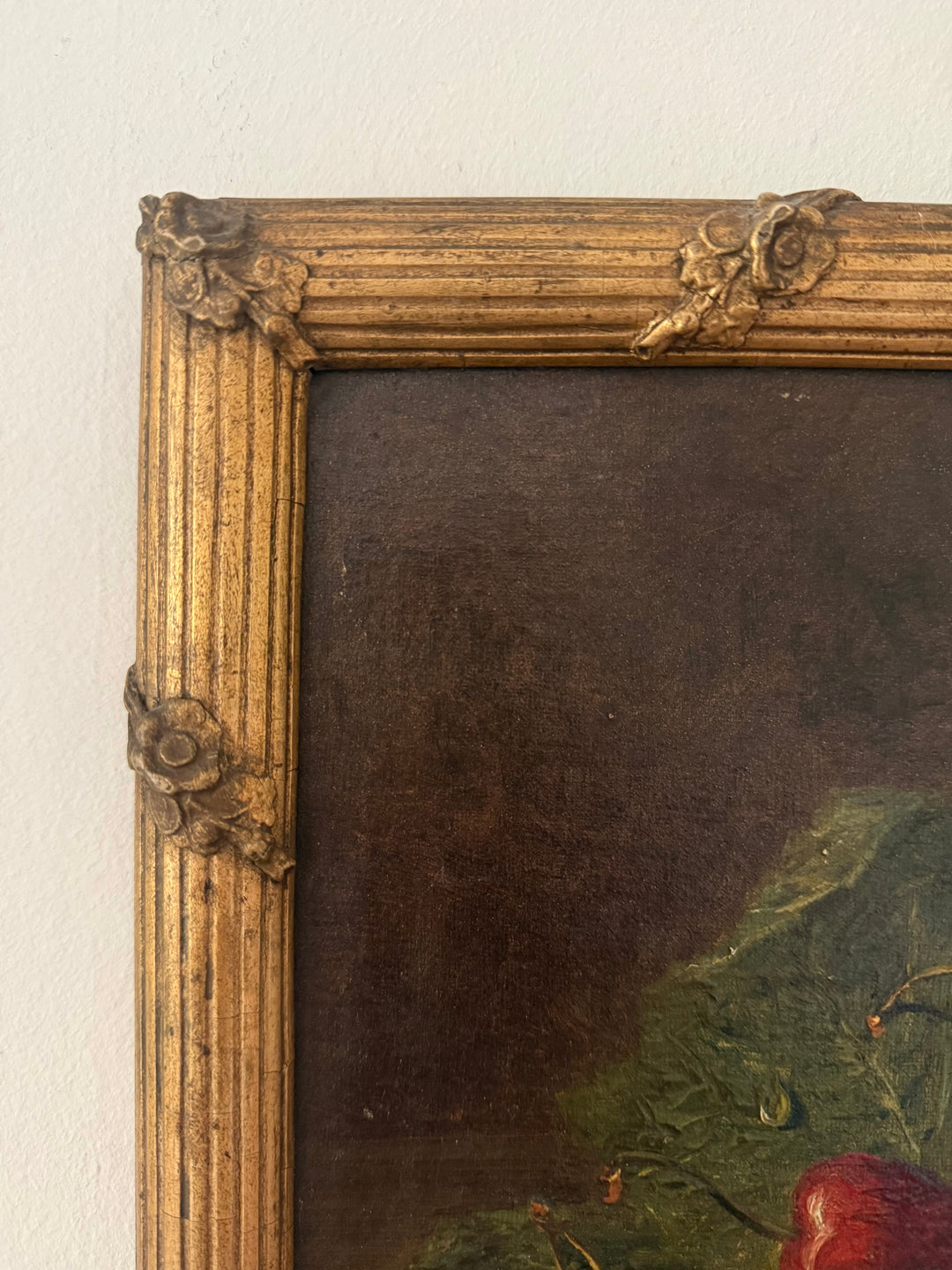Close-up of an ornate gold gilded picture frame with carved floral corner details, showing part of a dark painted canvas and a small portion of a still-life artwork.