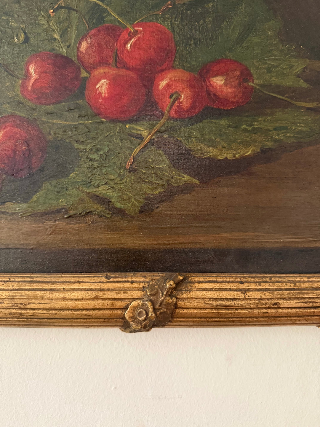 Close-up of a still life painting depicting red cherries on green leaves, mounted on a wooden panel with a decorative metal accent above a white wall.