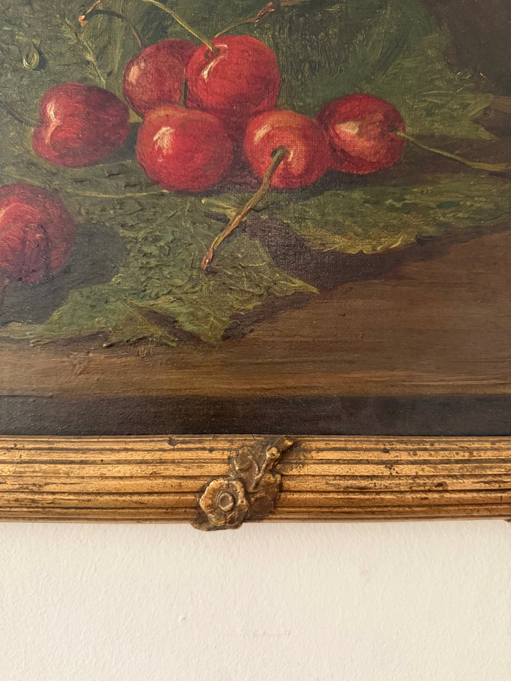 Close-up of a still life painting depicting red cherries on green leaves, mounted on a wooden panel with a decorative metal accent above a white wall.
