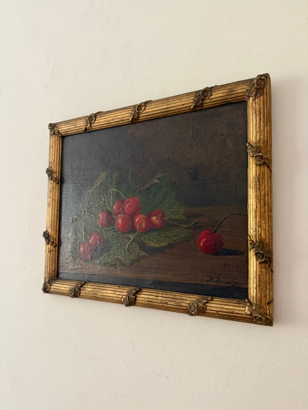 A framed still life oil painting depicting red cherries on green leaves against a dark background, housed in an ornate gold-colored frame and mounted on a plain off-white wall.
