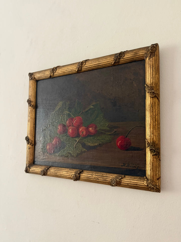 A framed still life oil painting depicting red cherries on green leaves against a dark background, housed in an ornate gold-colored frame and mounted on a plain off-white wall.