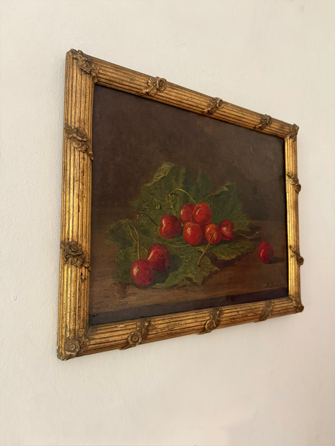 A framed antique still life oil painting depicting red cherries on green leaves, set in an ornate gold-colored frame and mounted on a plain white wall by Hubert Bellis 