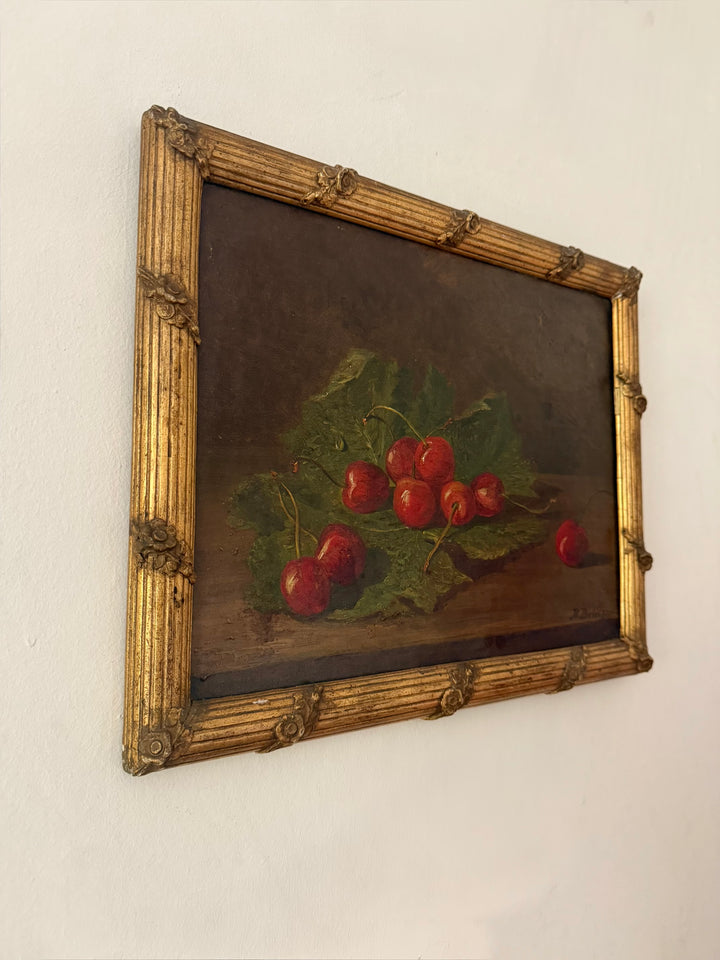 A framed antique still life oil painting depicting red cherries on green leaves, set in an ornate gold-colored frame and mounted on a plain white wall by Hubert Bellis 