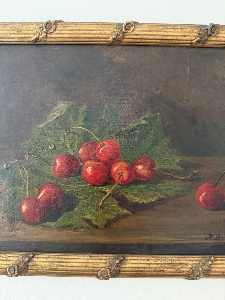 A framed still life oil painting depicting red cherries on green leaves, set against a dark brown background, enclosed in a decorative gold-toned frame.