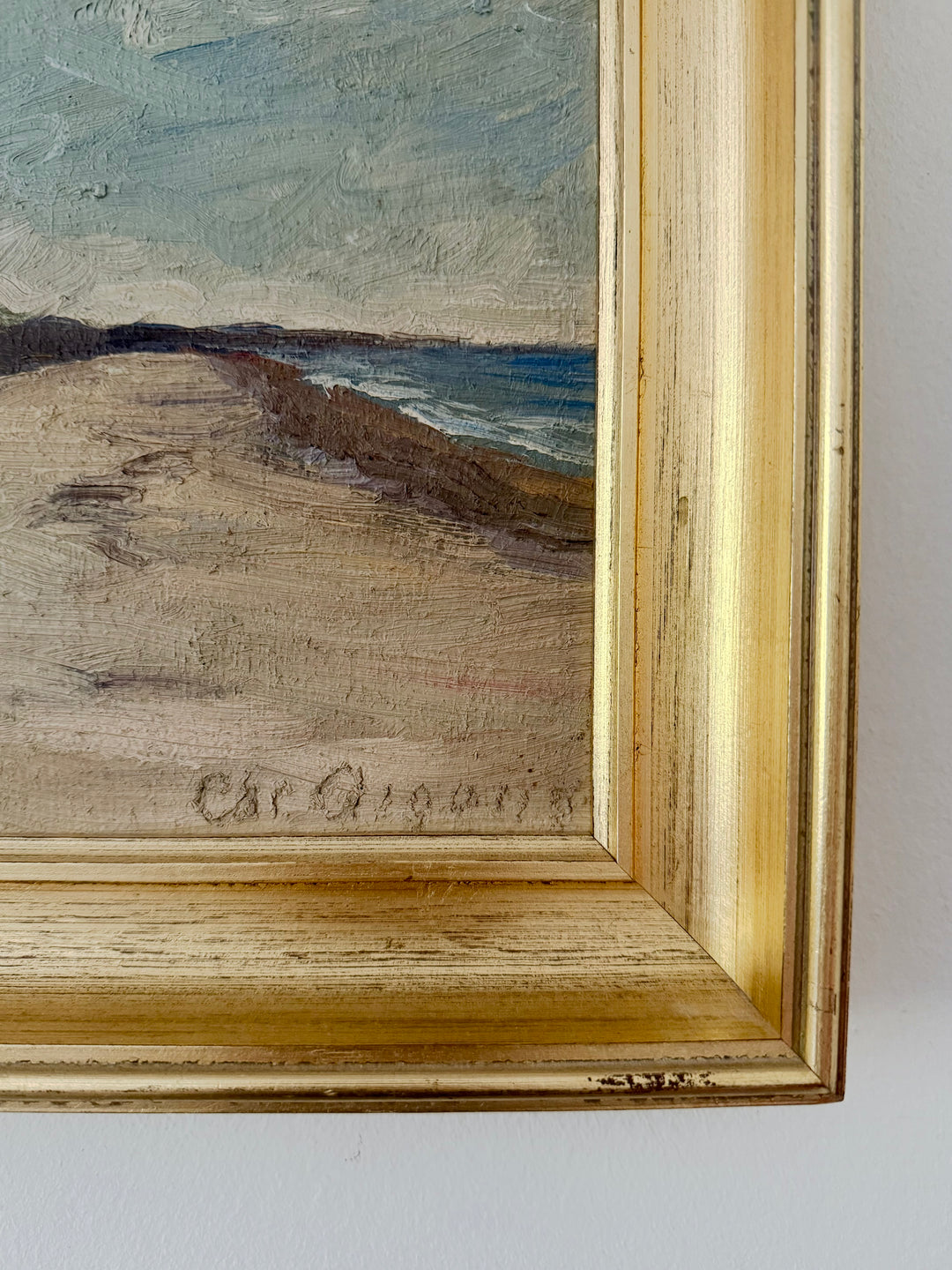 Close-up of a framed beach landscape painting with visible brushstrokes, showing sand, water, and sky, bordered by a distressed gold frame against a white wall.