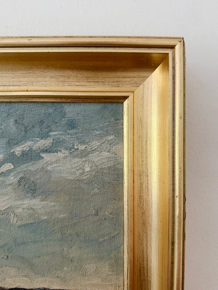 Close-up of a framed painting showing a textured blue-gray landscape or seascape with thick impasto brushstrokes, set in a distressed gold frame against a light neutral wall.