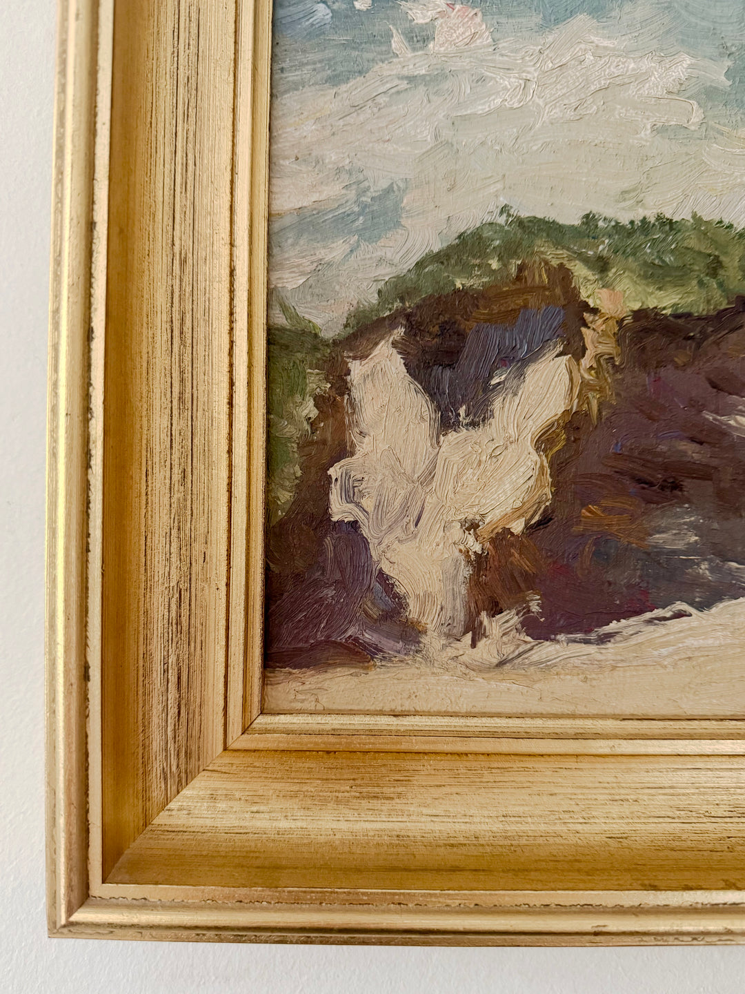Close-up of a framed landscape painting showing textured brushstrokes of white cliffs, green hills, and a cloudy sky, set in a light natural wood frame.
