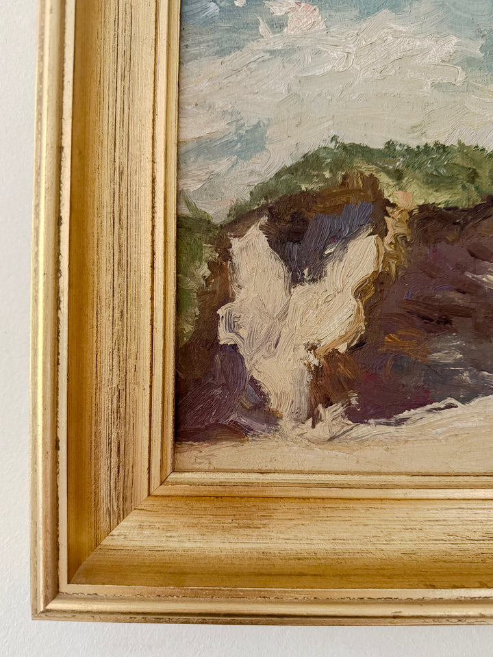 Close-up of a framed landscape painting showing textured brushstrokes of white cliffs, green hills, and a cloudy sky, set in a light natural wood frame.